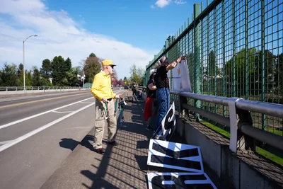 Activists from the Indivisible movement unfurl protest banners along the green mesh fencing of a North Portland overpass, their message advocating for a May 1st General Strike visible to Interstate 5 traffic below. A man in a bright yellow jacket and cap observes as his companion secures white banners bearing bold black text against the afternoon sky. Spring sunlight casts sharp shadows across the concrete walkway, while suburban rooftops and mature trees frame this moment of civic engagement above the busy thoroughfare.