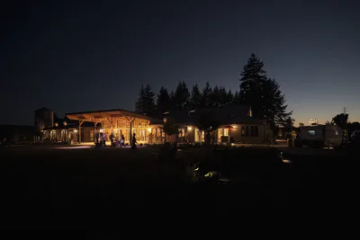 Golden light spills from the windows and covered porches of Abbey Road Farm's rustic buildings as twilight settles over Carlton, Oregon. Silhouettes of guests move across the illuminated terraces while towering evergreen trees frame the pastoral scene against a deep indigo sky. The warm architectural lighting creates an inviting atmosphere that contrasts beautifully with the surrounding darkness of the Willamette Valley countryside.