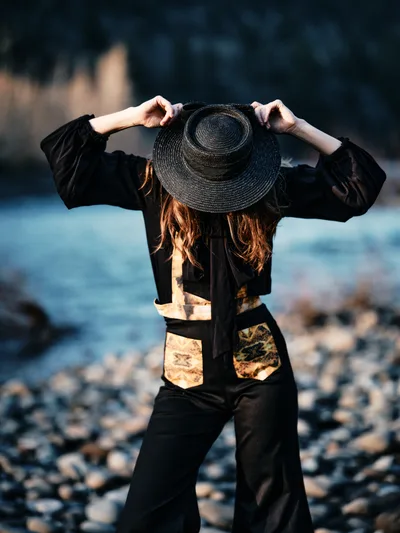 A solitary figure in black clothing adjusts her wide-brimmed straw hat while standing on the rocky riverbed of Oregon's Columbia River Gorge. Golden afternoon light illuminates her auburn hair and creates warm patches on her dark jacket, while the blurred water and distant forested hills provide a serene backdrop. The composition captures a moment of quiet introspection along the Floodplain Trail in Multnomah County.