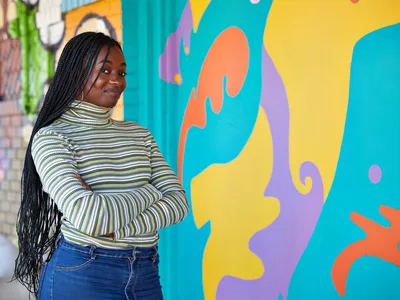 Naomi Likayi stands confidently with crossed arms before her vibrant mural in Portland's Alberta Arts District. The young Black artist wears a striped turtleneck and denim, her long braids cascading as she poses against the flowing abstract forms painted in brilliant turquoise, coral, lavender, and sunshine yellow. The colorful street art transforms the urban wall into a celebration of contemporary African American artistic expression in Northeast Portland.