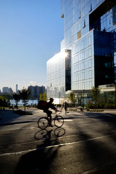 A silhouetted cyclist glides through the dappled shadows of Kent Avenue as late afternoon sun blazes between towering glass residential buildings along Brooklyn's East River waterfront. The dramatic backlighting creates sharp geometric patterns on the pavement while the gleaming facades of modern high-rises reflect the golden hour radiance. Pedestrians stroll leisurely in the background, framed by manicured landscaping and the distant Manhattan skyline across the water.