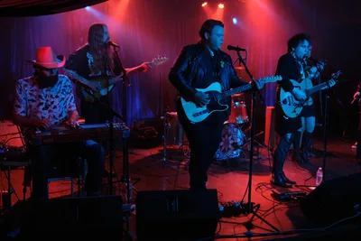 The four-piece band Roselit Bone commands the intimate stage at Dante's in Portland's music scene, bathed in dramatic red and blue theatrical lighting. Barry Walker mans the keyboards while wearing a distinctive cowboy hat and patterned shirt, as Tobias Berblinger anchors the rhythm section on bass. Victor Franco delivers vocals while wielding his electric guitar center stage in a black leather jacket, with Charlotte McCaslin adding layers on guitar from stage right. The moody lighting transforms the cramped venue into an atmospheric sonic cathedral, cables snaking across the floor between amplifiers and effects pedals.