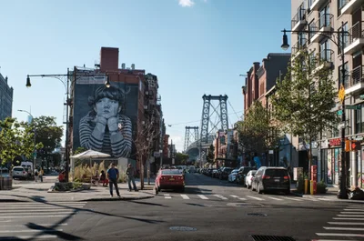 A striking black and white mural of a contemplative child dominates the brick facade of a building on Bedford Avenue in Williamsburg, Brooklyn. The monochromatic portrait creates a powerful contrast against the warm afternoon light that bathes the bustling street scene, where pedestrians cross the intersection as traffic flows toward the iconic Manhattan Bridge towers visible in the distance. The composition captures the vibrant street life of this gentrified neighborhood, where contemporary residential buildings with modern balconies stand alongside older brick structures adorned with large-scale street art.