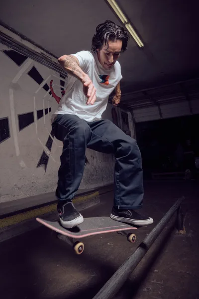 A focused skateboarder with tousled dark hair and visible tattoos navigates the gritty confines of Commonwealth Skateboarding's indoor bowl in Portland's Buckman neighborhood. Fluorescent lighting cuts through the shadowed concrete terrain, illuminating his white graphic tee and baggy jeans as he maintains perfect balance on his deck. The raw industrial atmosphere of the Southeast Portland skate shop creates an authentic backdrop for this intimate editorial moment.