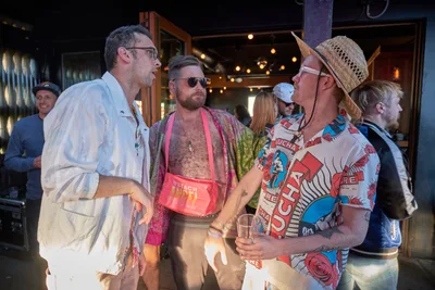 Three men in vibrant summer attire engage in animated conversation during Your Sunday Best's season opener at White Owl Social Club in Portland. The scene captures the eclectic spirit of the daytime party, with one guest sporting a pink mesh tank and bandana, another in a woven hat and patterned shirt, while warm Edison bulb lighting creates an intimate atmosphere against the venue's industrial backdrop. The candid moment embodies the relaxed social energy of Portland's alternative summer scene.