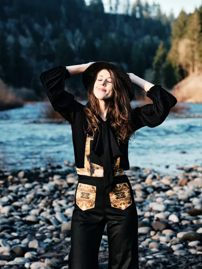 A young woman with flowing chestnut hair stands in peaceful contemplation along Oregon's Floodplain Trail, her arms gracefully raised to adjust her black wide-brimmed hat. She wears a distinctive black ensemble adorned with golden leaf-pattern accents that echo the autumn landscape behind her. The rocky riverbed of what appears to be the Sandy River stretches beneath her feet, while soft natural light illuminates her serene expression against the backdrop of dense Pacific Northwest forest.
