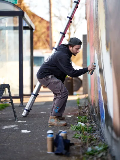 Travis Fields, known as campographic, works intently on a vibrant mural depicting poet Amanda Gorman at the intersection of Northeast 15th Avenue and Killingsworth Street in Portland's Alberta district. The bearded artist in dark hoodie and backwards cap stretches toward the wall with spray can in hand, his extension ladder propped nearby. Scattered paint cans dot the weathered pavement while soft afternoon light illuminates this urban canvas transformation in one of Portland's culturally rich neighborhoods.