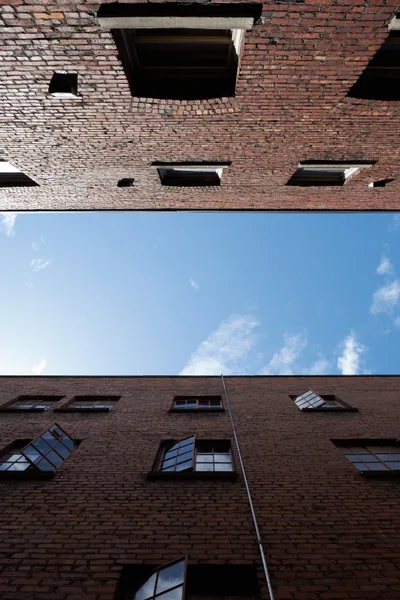 A dramatic worm's-eye view captures the towering brick walls of a historic warehouse courtyard in Portland's Central Eastside Industrial District. The weathered red brick facades frame a narrow slice of azure sky dotted with white clouds, while industrial windows reflect the afternoon light. The geometric composition emphasizes the imposing height and industrial heritage of this converted warehouse space on Southeast Yamhill Street.
