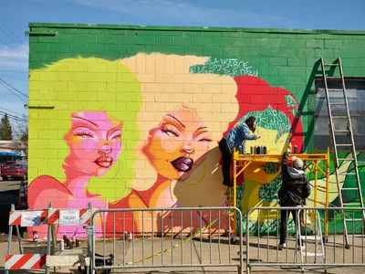 Multidisciplinary artist Vilatose works from scaffolding to paint a vibrant pop art mural titled 'Jardín Cubana' featuring three female figures on the green brick wall of a building in Portland's Alberta Arts District. The artist applies paint with careful precision while safety barriers protect the work zone below, capturing the creative energy of Northeast Alberta Street's thriving street art scene under clear blue skies.