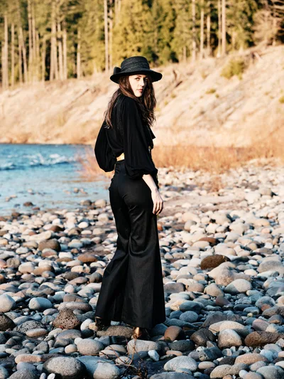 A woman in flowing black attire and wide-brimmed hat stands contemplatively among the weathered river stones of Oregon's Floodplain Trail. Golden hour light bathes the pebbled riverbed and distant forested hillsides in warm amber tones, creating an atmosphere of quiet introspection. Her silhouette against the natural landscape evokes a sense of solitary wandering and connection with the Pacific Northwest wilderness.