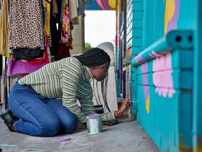 Naomi Likayi kneels on the sidewalk outside a vibrant clothing boutique, carefully signing her completed mural on turquoise-painted brick walls. Her striped long-sleeve shirt and concentrated expression capture the intimate moment of artistic completion in Portland's Alberta Arts District. Colorful fabrics and garments hang in the shop window behind her, creating a rich tapestry of community commerce and creative expression.