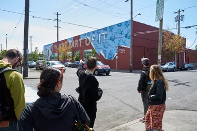 A diverse group of participants gathers on Southeast Taylor Street in Portland's Central Eastside district, their attention focused on a vibrant mural adorning a red brick industrial building. The afternoon sun illuminates the scene as tour guide Tomás Valladares leads an interpretive discussion about the neighborhood's street art culture. Power lines create geometric patterns overhead while parked cars and urban infrastructure frame this moment of cultural discovery and community engagement.