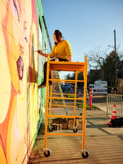 Multidisciplinary artist Villatose works from an orange rolling scaffold against the brilliantly colored mural 'Jardín Cubana' on Northeast Alberta Street in Portland's Alberta Arts District. Golden hour light bathes the scene as the artist, wearing a yellow sweater, applies brushstrokes to the vivid composition featuring three female figures in green, pink, and yellow. Traffic cones and street barriers frame the urban workspace, while bare winter trees and power lines create geometric patterns against the clear blue sky.