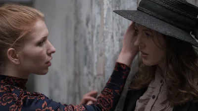 A close, intimate portrait of two women facing each other against a weathered concrete wall. Faith Yakovleva, seen in profile on the left, wears a dark floral top with red and black flowers and has her strawberry blonde hair pulled back. She reaches out to gently touch the brim of a wide black straw hat worn by Rhianna Atwood, who stands against the wall on the right in a cream ruffled blouse, her eyes downcast.