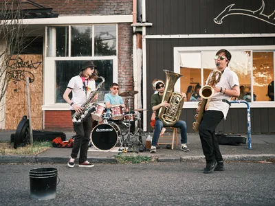 A spirited brass quartet brings jazz to the sidewalk during Last Thursday's street fair on Alberta Street in Portland's Concordia neighborhood. Two saxophonists flank the ensemble while a tuba player and drummer anchor the rhythm section, their instruments gleaming in the late afternoon light. The urban backdrop of weathered brick buildings and storefront windows frames this impromptu concert, capturing the authentic spirit of Portland's monthly art walk where music spills onto the pavement and community gathers around creativity.