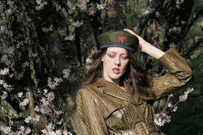 Model Rhianna Atwood poses contemplatively among blooming cherry blossoms at Mount Tabor in Portland, Oregon, wearing a golden crocodile-textured leather coat and olive military cap adorned with a red star. The soft spring light filters through the delicate pink sakura petals, creating an evocative juxtaposition between militant iconography and nature's gentle renewal. Her hand rests thoughtfully against the cap's brim as she gazes downward, surrounded by the ephemeral beauty of hanami season.
