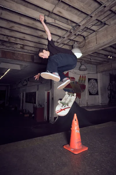 A skateboarder executes a dynamic jump over a weathered orange traffic cone inside Commonwealth Skateboarding in Portland's Buckman neighborhood. The dramatic low-angle perspective captures the athlete mid-flight against the industrial basement ceiling, with exposed beams and graffiti-adorned concrete walls creating a raw urban backdrop. The skater's concentrated expression and athletic form demonstrate the precision required for this technical maneuver. Harsh artificial lighting casts deep shadows across the underground skate space, emphasizing the gritty authenticity of Portland's skateboarding culture.