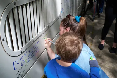 A TriMet train operator in a blue scrunchie and denim jacket leans over her young child in a royal blue sweatshirt as they write farewell messages on the white exterior of a MAX Type 1 train car during its retirement ceremony at Holladay Park in Portland's Lloyd District. Both concentrate intently as they add their handwritten notes to the growing collection of colorful marker messages covering the train's surface. The circular ventilation grate and industrial design of the vintage transit car frame their tender moment of personal connection to the retiring fleet.