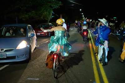 Under the glow of streetlights on Southeast Lincoln Street in Portland's Richmond neighborhood, costumed cyclists participate in the Drop Out Prom Ride during Pedalpalooza 2019. A cyclist in an elaborate turquoise ball gown and yellow helmet weaves between cars on the wet asphalt, while another rider in blue Victorian attire follows nearby. The nocturnal procession transforms the ordinary street into a theatrical stage, where formal wear meets bicycle culture in Portland's celebrated cycling festival.
