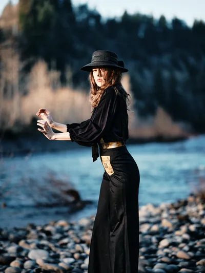 A woman in flowing black attire and wide-brimmed hat stands in contemplative pose along the rocky shoreline of the Columbia River Gorge. Golden hour light catches her profile as she cradles a glass bottle, creating an intimate moment against the backdrop of Oregon's forested riverbank. The composition balances her silhouetted form against the soft blur of water and distant treeline, evoking themes of solitude and reflection in nature.