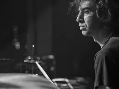 Julian Dorio of Band of Skulls sits in quiet contemplation behind his drum kit at the legendary Webster Hall in Manhattan during the band's 10 Year Anniversary Tour. The black and white portrait captures the drummer in profile against a backdrop of soft stage lights, with drumsticks poised in his hands. The atmospheric lighting creates dramatic shadows across his face while the blurred venue lights in the background suggest the intimate energy of this historic East Village music venue.