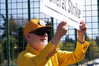 A determined activist in a bright yellow shirt and baseball cap holds aloft a white banner reading "General Strike" during a protest action near North Portland's Interstate 5 overpass. The man's weathered face shows quiet resolve as golden afternoon sunlight illuminates the scene, while chain-link fencing and urban infrastructure frame the background. His raised arms and focused expression capture the earnest spirit of grassroots political organizing in the Pacific Northwest.