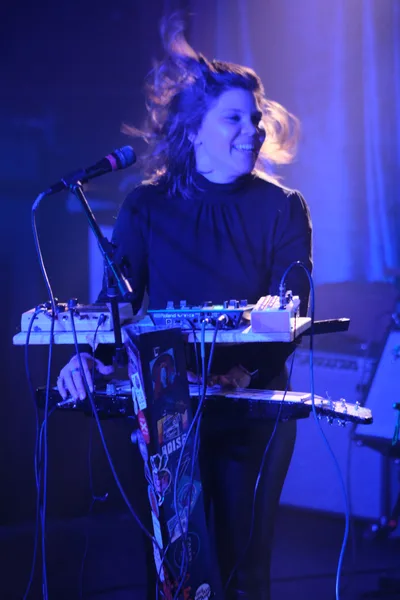 A female electronic musician from experimental band Sun Blood Stories performs behind a Roland synthesizer setup at Dante's venue in downtown Portland, her blonde hair catching movement as she leans toward the microphone. Saturated blue and purple stage lighting bathes the intimate performance space, creating an otherworldly atmosphere that transforms the musician and her equipment into silhouettes against vertical light columns. The industrial venue's brick walls dissolve into darkness beyond the concentrated beam lighting, while cables snake across the stage connecting her multi-tiered keyboard rig.