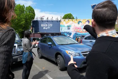 Tour guide Tomás Valladares gestures enthusiastically toward vibrant murals adorning the River City Bicycles building as visitors gather in the bustling Central Eastside district. The afternoon scene captures the intersection of urban art appreciation and community engagement, with participants framed against colorful street art featuring stylized figures. Shadows stretch across the pavement while bicycles crown the rooftop installation, creating a quintessentially Portland moment where cycling culture meets artistic expression.