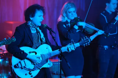 Charlotte McCaslin commands the foreground with her pristine white hollow-body guitar, singing into the microphone while bathed in electric blue stage lighting. Behind her, Faith Grossnicklaus draws her violin bow across the strings, both musicians absorbed in their performance at the intimate Dante's venue. The atmospheric lighting creates a dreamlike quality, with deep purples and blues washing over the duo as they deliver their ethereal sound to the RX Fest 2019 audience. The composition captures the intimate yet electric energy of live performance in Portland's renowned underground music scene.