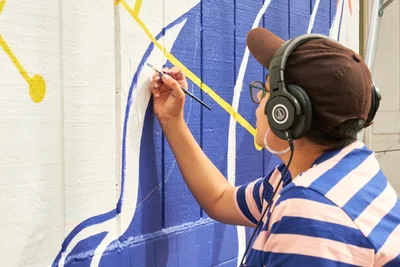 Maria Rodriguez, known as Sparkykneecap, works intently on her vibrant mural "Let's Talk" at Open Signal in Portland's Eliot neighborhood. Wearing headphones and a blue-striped shirt, the Mexican-American artist carefully applies precise brushstrokes to the bold geometric design featuring brilliant blues and yellows against white brick. The concentrated creative process unfolds in natural daylight, capturing the meditative focus required for large-scale public art.
