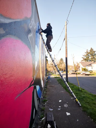 Street artist Travis Fields, known as campographic, climbs a silver extension ladder to work on a vibrant mural of poet Amanda Gorman in Portland's Alberta neighborhood. The afternoon light illuminates swirling pink, coral, and blue paint across the building's white facade as the artist works methodically on the large-scale tribute. Power lines cross the clear sky above this residential intersection, while scattered paint remnants on the weathered sidewalk below tell the story of artistic creation in progress.