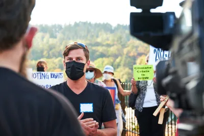 Michael Gaskill, wearing a black face mask and v-neck shirt, stands composed as he speaks to a KOIN Channel 6 news reporter during a climate change demonstration outside Representative Kurt Schrader's office in Oregon City. Behind him, fellow protesters hold signs including "Climate Crisis Is NOW" and "VOTING RIGHTS" against the backdrop of Oregon's rolling forested hills. The scene captures the intersection of grassroots activism and media coverage in the afternoon light of McLoughlin Promenade.