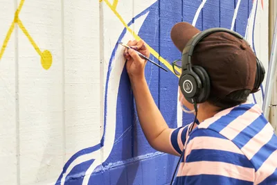 Mexican-American artist Maria Rodriguez, known as Sparkykneecap, meticulously details a vibrant blue and yellow mural at Open Signal in Portland's Eliot neighborhood. Wearing black headphones and a blue-striped shirt, they work with focused concentration on the community-commissioned piece titled 'Let's Talk.' The afternoon light illuminates their precise brushwork against the bold geometric patterns that explore themes of identity and cultural connection.