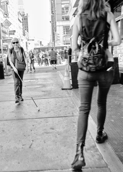 A striking black and white street photograph captures two figures moving in opposite directions along the bustling sidewalks of Chelsea's West 23rd Street. In the sharp foreground, a woman with flowing hair strides purposefully past the camera, while in the softly focused background, a blonde woman wearing sunglasses and carrying a white cane navigates the urban landscape with quiet determination. The dramatic contrast between the crystal-clear foreground subject and the deliberately blurred background creates a powerful visual metaphor for the different ways we all move through the city's relentless rhythm.