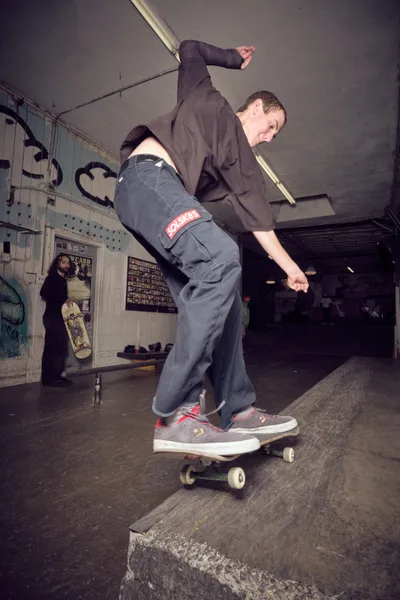 A skateboarder in dark casual wear and gray pants carves through the weathered concrete transitions of Portland's Commonwealth Skateboarding shop. The industrial interior buzzes with underground energy, graffitied walls and skateboard memorabilia creating an authentic backdrop. Fluorescent lighting casts dramatic shadows as the rider flows through the bowl with focused intensity, embodying the raw spirit of Pacific Northwest skate culture.