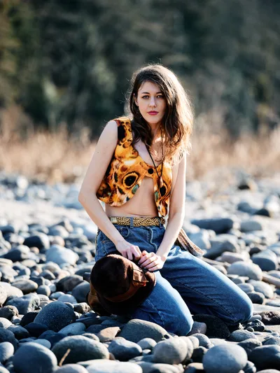 A young woman with flowing brown hair kneels gracefully among smooth river stones along Oregon's Floodplain Trail in Multnomah County. She wears a vibrant orange patterned crop top and denim jeans cinched with a woven belt, her pose contemplative against the sun-dappled backdrop of golden grassland and distant forest. The warm afternoon light creates a natural rim lighting effect, highlighting her figure against the softly blurred woodland setting.