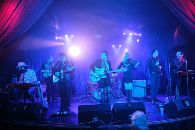 Seven musicians command the intimate stage at Dante's in Portland, bathed in electric blue stage lighting that cuts through veils of atmospheric haze. The band Roselit Bone performs beneath dramatic crimson curtains that frame the venue's signature cabaret atmosphere, their silhouettes emerging from the smoky darkness as spotlights pierce the ethereal performance space. Guitars, violin, and keyboards blend in this underground music sanctuary where Portland's alternative scene converges.