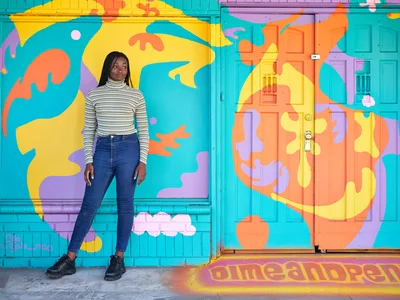 Artist Naomi Likayi stands beside her completed mural on Northeast 15th Avenue in Portland's Alberta district, her hand touching the vibrant wall art she has just signed. The explosive composition features flowing organic shapes in turquoise, coral, yellow, and purple that transform the building's facade into a celebration of color and movement. Wearing a striped turtleneck and dark jeans, Likayi poses confidently against her creation, which incorporates whimsical elements like floating clouds and abstract forms that seem to dance across the architectural surface.