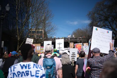 Demonstrators gather beneath Portland's historic Burnside Bridge in the Old Town district, their handmade signs creating a forest of dissent against azure spring skies. The crowd flows along Southwest Ankeny Street, with protesters of all ages holding placards declaring "LET'S CHANGE" and "IF WE ARE CAPABLE" among other messages of resistance. Bare-branched trees frame the scene while onlookers line the bridge above, witnessing this grassroots political mobilization in the heart of Portland's Chinatown neighborhood.