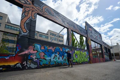 A guide from the Portland Street Art Alliance leads an interpretive tour beneath a towering industrial bridge adorned with vibrant graffiti murals in Portland's Hosford-Abernethy neighborhood. The scene captures the intersection of urban decay and creative renewal, where colorful street art transforms concrete infrastructure into an outdoor gallery. Dramatic clouds frame the composition, highlighting the contrast between the gritty industrial setting and the dynamic artistic expression that covers every available surface.