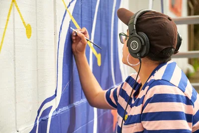 Mexican-American artist Maria Rodriguez, known as Sparkykneecap, works intently on her vibrant blue and yellow mural titled "Let's Talk" at Open Signal in Portland's Eliot neighborhood. Wearing headphones and a brown cap, she applies precise brushstrokes to the wall while dressed in a blue and white striped shirt. The afternoon light illuminates her focused expression as she brings themes of identity and cultural connection to life through bold geometric forms and warm colors.