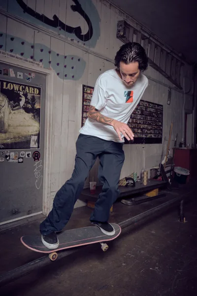 A concentrated skater navigates his board through  Commonwealth Skateboarding in Portland's Buckman neighborhood. Dramatic overhead lighting cuts through the dim space, illuminating vintage skate posters and memorabilia that plaster the weathered walls. The intimate setting captures the raw authenticity of underground skate culture, where passion thrives in unconventional spaces.