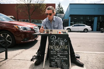 Till Gwinn, a local street poet, sits with legs spread apart on the sidewalk along Northeast Alberta Street in Portland, Oregon, crafting personalized poetry for passersby. Dressed in a light blue button-down shirt and dark sunglasses, he works intently at a small folding table adorned with a chalkboard sign advertising "Poetry of Your Prerogative Topics." The urban scene unfolds behind him with parked cars, brick buildings, and the characteristic overcast Pacific Northwest sky filtering soft, even light across the pavement.
