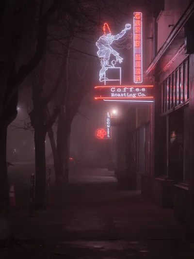 The iconic Caffe Vita Coffee Roasting Company neon sign pierces through a heavy fog on Alberta Street in Portland's Concordia neighborhood. The vintage-style sign features a whimsical figure holding a coffee cup, rendered in electric blue and white neon, while red lettering spells out the cafe's name below. Bare winter tree branches frame the left side of the composition as the atmospheric haze transforms the nighttime street into a dreamlike urban tableau.