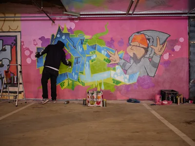 A black-clad artist works intently on a vibrant mural featuring dimensional blue and green lettering alongside a bearded character throwing up peace signs, all set against a hot pink backdrop in Clay Creative's parking garage. The industrial space comes alive with creative energy as spray paint cans and art supplies scatter across the concrete floor. Exposed ceiling elements and raw concrete walls frame this underground artistic sanctuary in Southeast Portland's Hosford-Abernethy neighborhood.