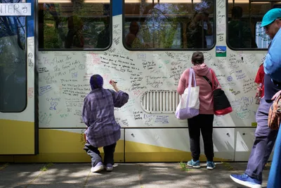 Commuters and transit enthusiasts gather around a retiring TriMet MAX Type 1 light rail car in Portland's Lloyd District, adding their handwritten messages and signatures to the vehicle's white exterior. A person in a plaid shirt crouches while writing on the train's side as others stand nearby with bags and belongings, creating an intimate farewell scene bathed in dappled afternoon sunlight. The train's windows reflect the surrounding urban trees, while countless personal messages in various inks cover nearly every inch of the accessible surface.