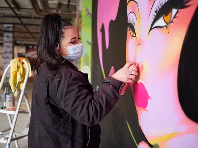 Multidisciplinary artist Vilatose, wearing a protective mask and dark coveralls, applies paint to a large-scale mural featuring a woman's face rendered in brilliant pinks and corals. Working within the industrial confines of Clay Creative's parking garage in Portland's Hosford-Abernethy neighborhood, she meticulously details the lips of her subject against the raw concrete backdrop. The contrast between the utilitarian space and the explosion of feminine color creates a striking juxtaposition of urban grit and artistic beauty.