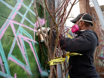 A hooded artist wearing a protective mask meticulously works on a vibrant mural at Clay Creative in Southeast Portland's Hosford-Abernethy neighborhood. The composition captures the intersection of nature and urban art as bare winter branches frame the scene, with spray paint cans suspended from the dormant tree. Soft daylight illuminates the artist's focused work against a backdrop of mint green and coral pink geometric patterns that dance across the weathered wall.