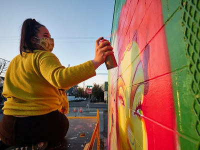 Multidisciplinary artist Vilatose, wearing a golden corduroy sweater and patterned face mask, spray paints bold coral and lime green hues onto her mural 'Jardín Cubana' featuring three female figures on a wall in Portland's Alberta Arts District. The late afternoon light bathes the scene in warm tones as she works methodically on the colorful mural, with urban street elements and traffic cones visible in the background. The composition captures the intimate moment of artistic creation against the backdrop of Northeast Portland's creative corridor.