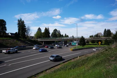 Activists from the Indivisible movement stage a banner drop from a pedestrian overpass spanning Interstate 5 in North Portland, their protest banner visible against the spring sky as afternoon traffic flows beneath. The concrete bridge arcs over multiple lanes of busy freeway traffic, with evergreen trees and residential neighborhoods framing the scene under wispy clouds. This moment captures civic engagement intersecting with daily commuter life, as the activists' May 1st General Strike message reaches drivers below on one of the Pacific Northwest's major transportation arteries.